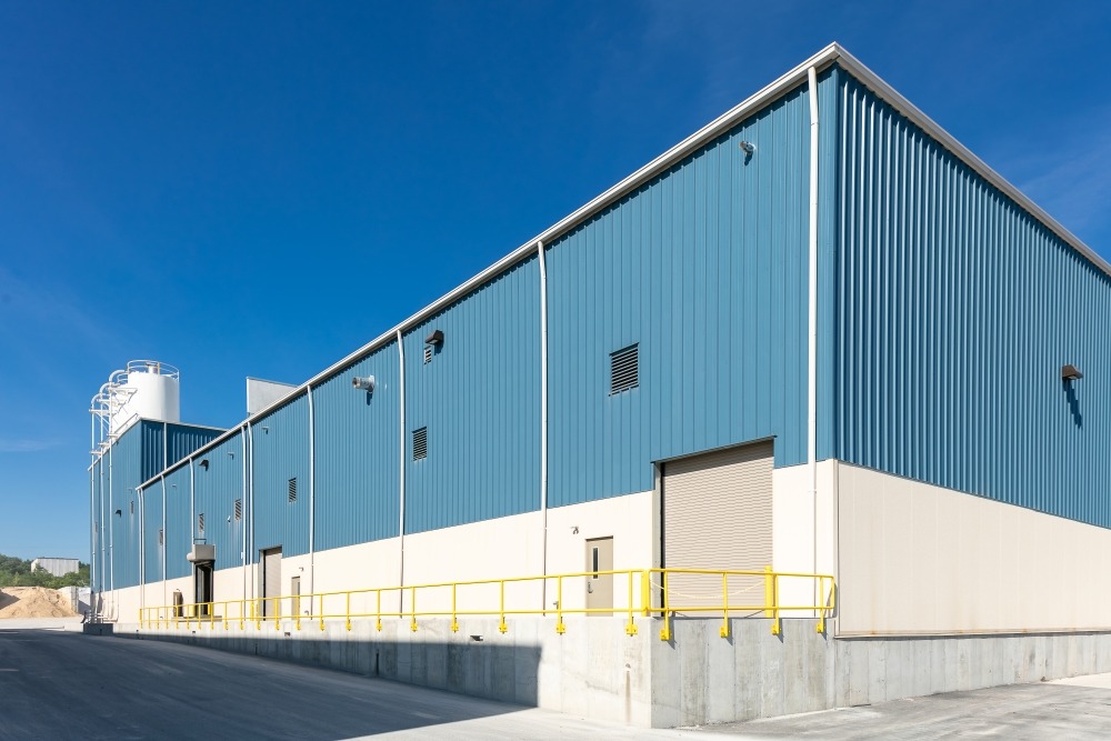 Blue and beige metal industrial building with loading dock, roll-up door, yellow safety railing, and rooftop ventilation equipment under a clear blue sky.